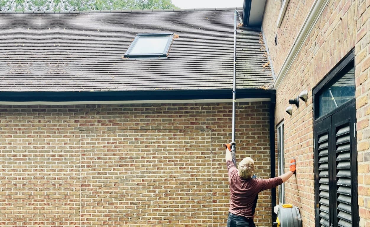 Person using a long telescopic pole to vacuum debris from high gutters on a brick home.